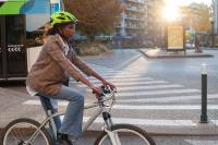 Black woman in a brown blazer, flared jeans, and a neon green helmet riding a gray bike through city streets.