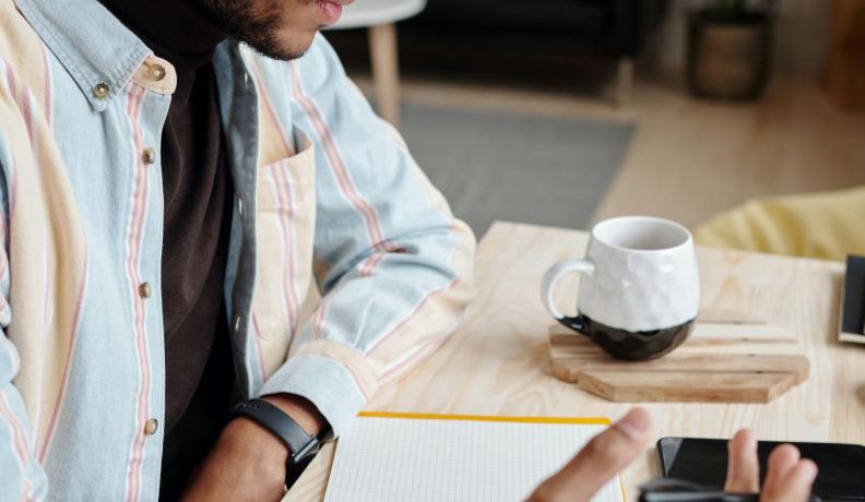 young black man wearing a flannel and studying or working at a desk with a notebook and a cup of coffee.