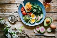 Plate of prepared veggies and boiled egg next to black pepper and sliced produce on a wooden table