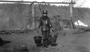 A black and white photo of a child under the age of 12 carrying two grease pails. He is covered in grease on his body. He stands alone in a bare and gray construction site on a hill.