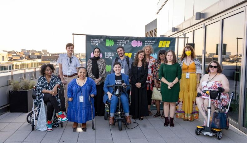 Twelve people of varying diversities and disabilities pose with canes, wheelchairs, and scooters in front of a Disability Culture Lab banner to celebrate the launch of the nonprofit.