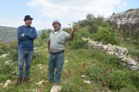 Nasser Abufarha stands to the left on a grassy hill in Palestine, arms crossed in a long-sleeve blue shirt and black hate. On the right is a local Palestinian farmer, in a grey shirt and turban, as they discuss regenerative agriculture.