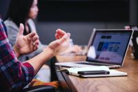 hands gesturing to explain something in a meeting. Computer and notebook are laid out on a table.