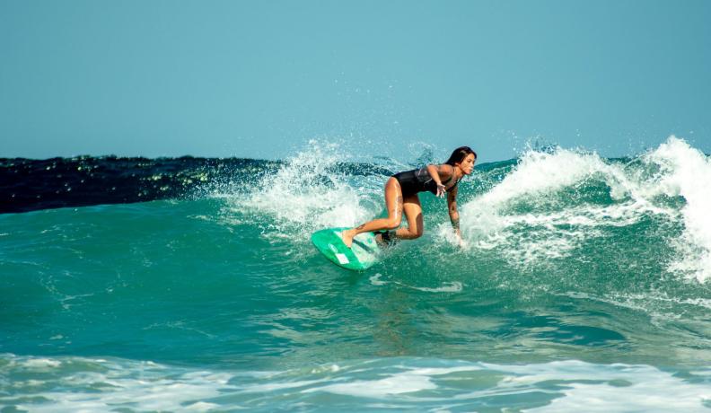 a filipina woman in a black one piece bathing suit rides the turquoise waves on her surfboard.