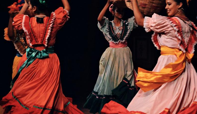 Women in traditional Mexican dresses dancing in a circle while holding baskets above their heads.