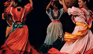 Women in traditional Mexican dresses dancing in a circle while holding baskets above their heads.