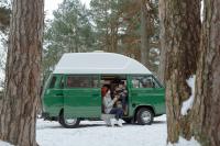 a couple sitting in their green van that they live out of. It is a snowy day and they are wearing sweaters and holding warm mugs, smiling at each other.