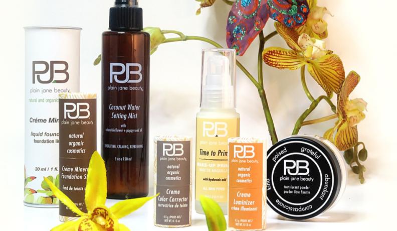 Plain Jane Beauty products in various tubes, spray bottles, and tubs, set against a white backdrop with flowers.