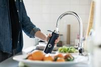 white person wearing a denim shirt and refilling a black metal water bottle in a kitchen sink.
