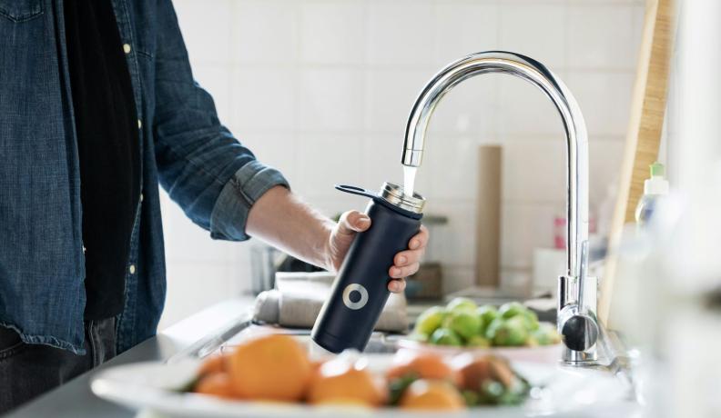 white person wearing a denim shirt and refilling a black metal water bottle in a kitchen sink.