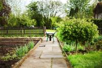 A wide shot of a wheelbarrow on a stone sidewalk. On the right are shrubs and grass, on the left is soil for a garden. Climate Victory Gardens.