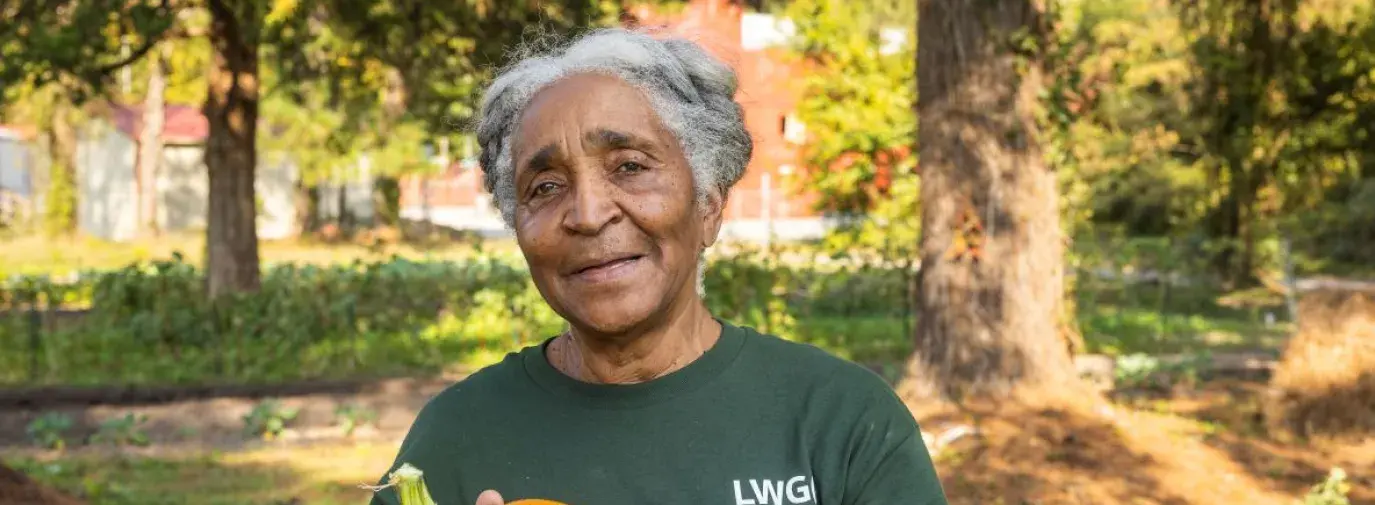 Cheryl Alston stands in a green long sleeve shirt holding a small pumpkin. She is looking calmly and pleasantly at the camera.