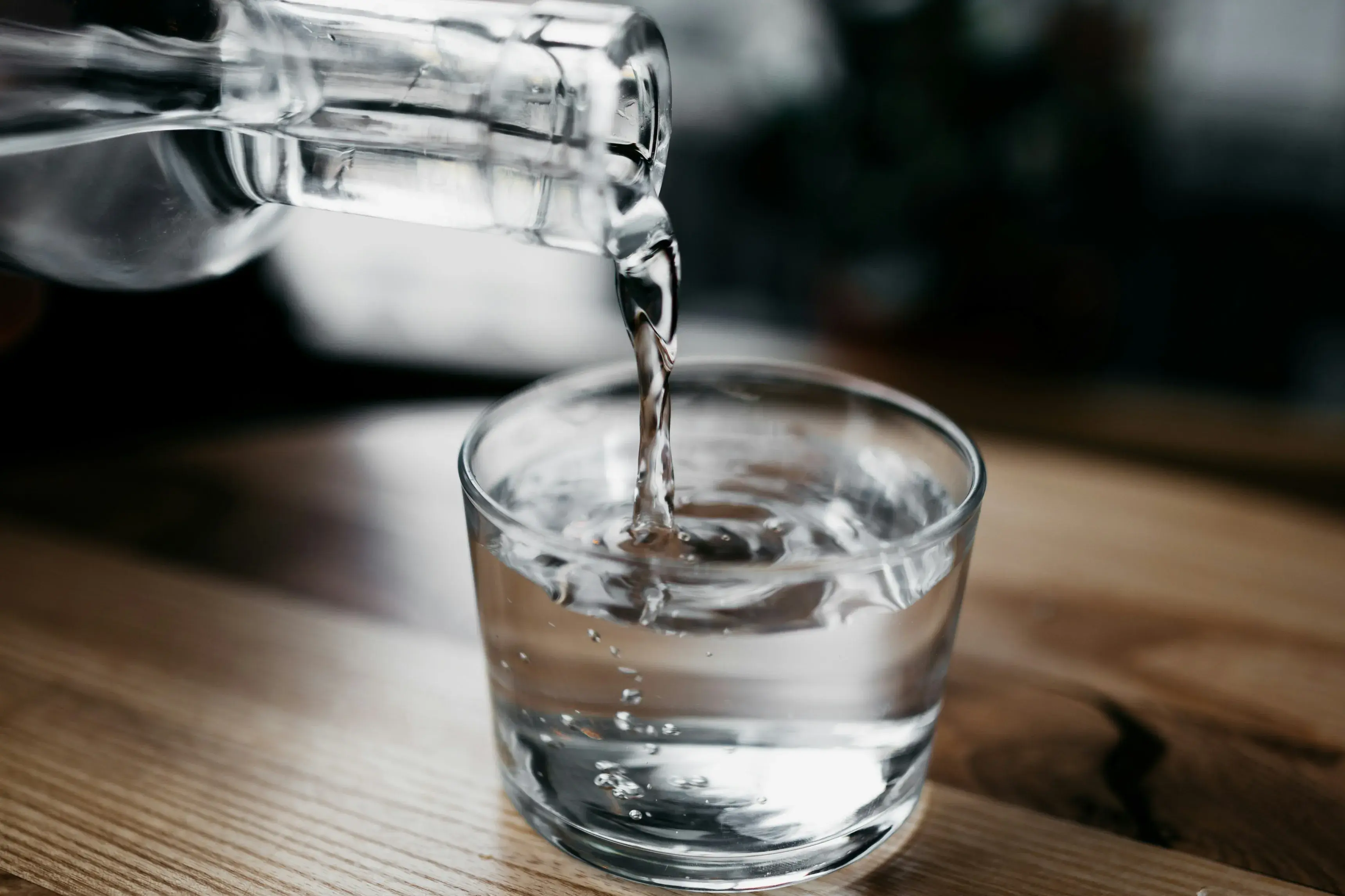 Water pouring from a glass bottle into a glass jar, which is set on a wooden table.