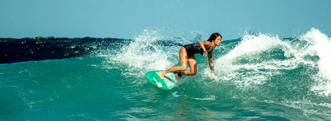 a filipina woman in a black one piece bathing suit rides the turquoise waves on her surfboard.