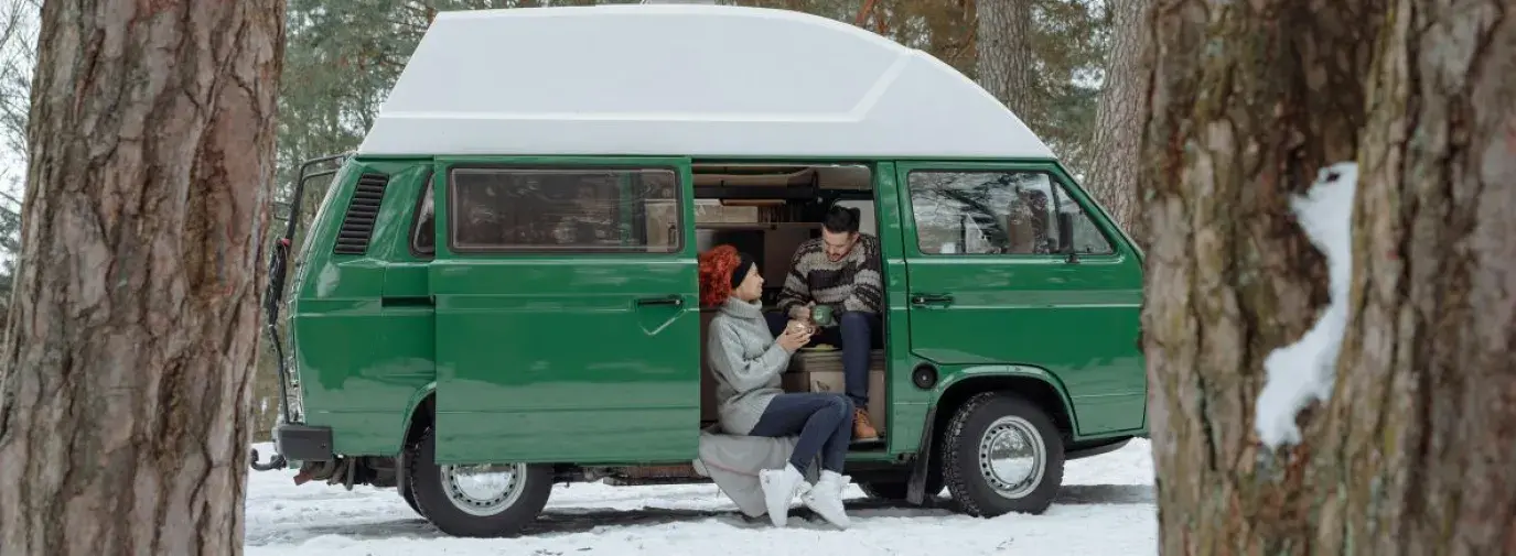 a couple sitting in their green van that they live out of. It is a snowy day and they are wearing sweaters and holding warm mugs, smiling at each other.