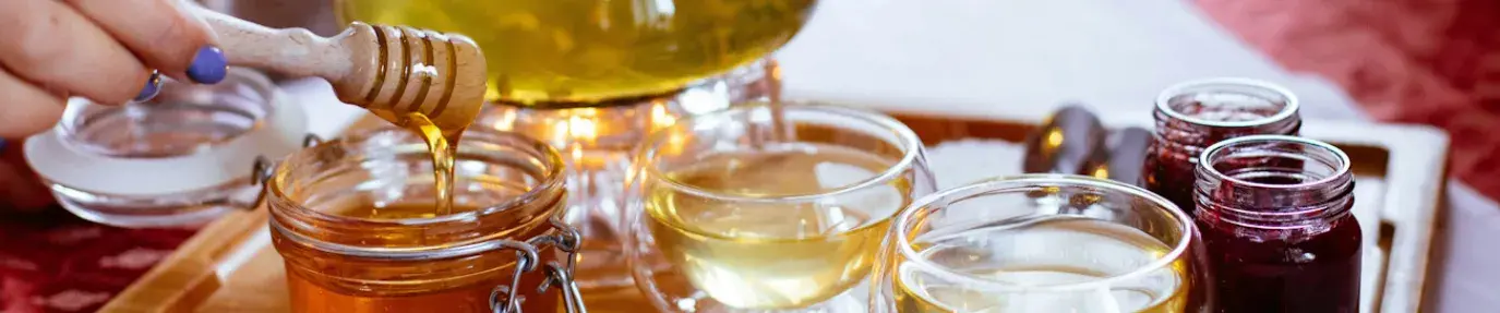 A wooden tray with tea in a glass kettle, jars of jam, and an open mason jar of honey. A hand sticks out from the left holding a honey dipper. Sustainable honey products